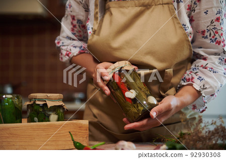 Close-up of the hands of a housewife in beige chef's apron, holding a glass jar of homemade pickled chili peppers. Marinating and preserving vegetables, healthy fermented food, winter natural products 92930308