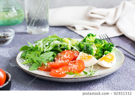 A plate with broccoli, tomato, arugula, boiled egg and sesame seeds on a plate on the table. 92931332