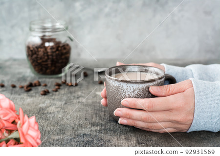 Latte in a ceramic cup in female hands, a jar of coffee beans and roses on the table. Latte in a ceramic cup in female hands, a jar of coffee beans and roses on the table. 92931569