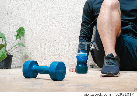 man sitting on the floor next to blue dumbbell and bottle with water, after exercising. exercise at home, white background. fitness concept. health concept, space for advertising text. 92931817