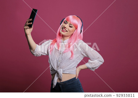 Young adult with pink hair taking pictures or recording video on smartphone, listening to music on headphones. Using mobile phone to take photos and enjoy songs on headset for entertainment. 92933811