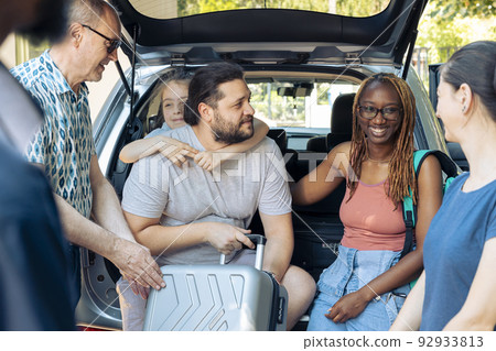 Diverse family and friends loading trolley in automobile trunk, preparing to leave on holiday trip. Travelling by car on summer vacation, going to seaside destination with multiethnic people. 92933813