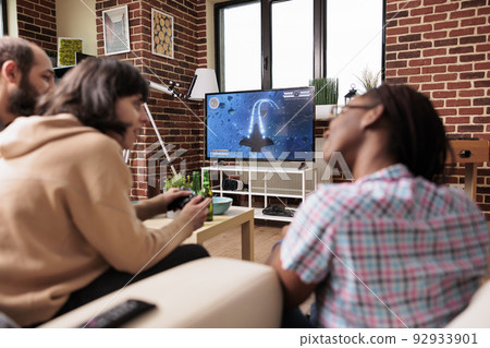 Multiethnic group of friends sitting at home in living room while playing space videogame simulator. People enjoying fun leisure activity on modern gaming console device while having beer and snacks. Multiethnic group of friends sitting at home in living room while playing space videogame simulator. People enjoying fun leisure activity on modern gaming console device while having beer and snacks. 92933901