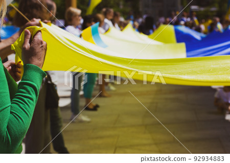 A crowd of patriots at rally in support of Ukrainians in Russia's war. People are holding a huge yellow blue Ukrainian flag, a symbol of Ukraine's independence. Save Ukraine in conflict. NATO summit. 92934883