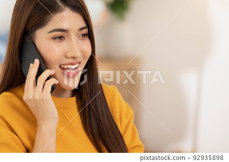 At home in the living room.Asian woman talks on the phone on the sofa in a calm attitude in the morning.Calling pals and having hanppy and good time. At home in the living room.Asian woman talks on the phone on the sofa in a calm attitude in the morning.Calling pals and having hanppy and good time. 92935898