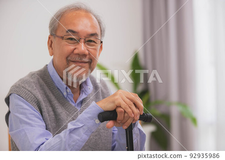 Asian Elderly man holding walking stick for physiotherapy.Sitting on sofa after walking in morning. Asian Elderly man holding walking stick for physiotherapy.Sitting on sofa after walking in morning. 92935986