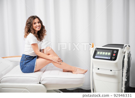 Cheerful young woman smiling at camera, sitting on daybed near diode laser epilation machine. Beautiful woman resting near hair removal device in cosmetology clinic. 92946331