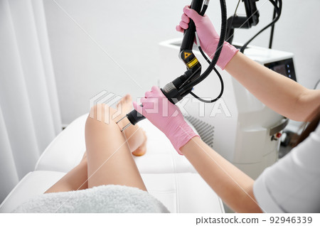 Close-up treatment of client's thigh with fractional CO2 laser. Laser skin ablation for subsequent scar treatment and local skin rejuvenation. Beautician performing modern laser resurfacing in salon. 92946339