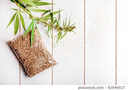 Transparent bag with cannabis seeds and a branch of fresh cannabis on a light wooden background Transparent bag with cannabis seeds and a branch of fresh cannabis on a light wooden background 92946917