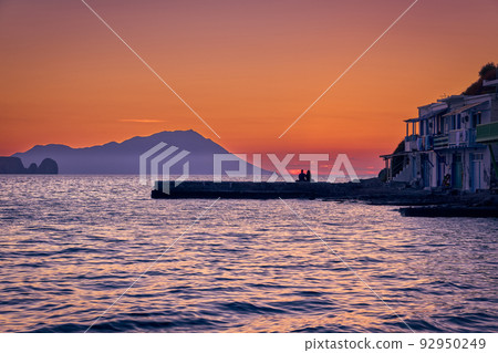 Sunset in fishermen village of Klima, Milos island, Greece. Pair of adults enjoy sunset view on pier of small fishermen's village. Hazy hills, dawn. Sunset in fishermen village of Klima, Milos island, Greece. Pair of adults enjoy sunset view on pier of small fishermen's village. Hazy hills, dawn. 92950249