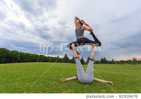 Man lying on grass and balancing woman in his feet. Young couple doing acro yoga in park 92950676