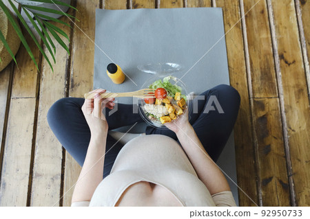 Young happy beautiful pregnant woman eating salad while sitting on yoga mat in lotus pose 92950733