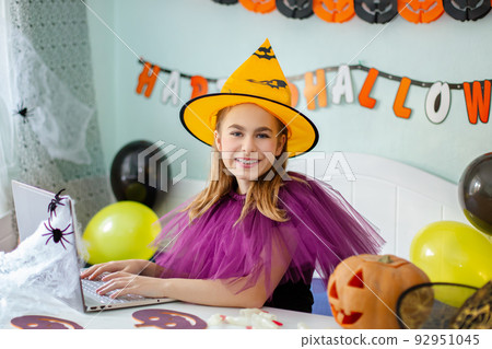 Cute little girl wearing witch hat sitting behind table Halloween theme decorated room. Preparation for the celebration. Cute little girl wearing witch hat sitting behind table Halloween theme decorated room. Preparation for the celebration. 92951045
