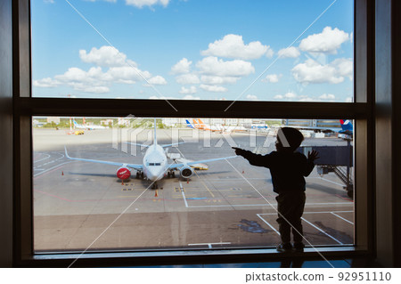 Moscow, Russia 07 14 22 Little boy carry bag waiting boarding to flight in airport transit hall and looking through the window at airplane departure. Active family lifestyle, travel by air with child  92951110
