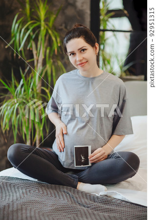 Pregnant woman holding pregnancy ultrasound while sitting in lotus pose on bed at home 92951315