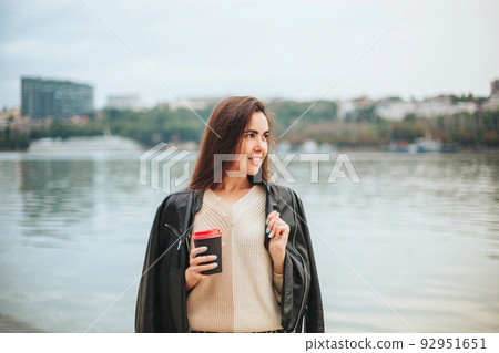 Young woman with take away coffee in hand in casual clothes near river water Young woman with take away coffee in hand in casual clothes near river water 92951651