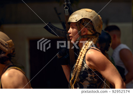 Caucasian Military lady woman in tactical gear posing for photo at summer season. Wearing green camo uniform and assault rifle, in military gear and headset, lady is looking at side 92952574