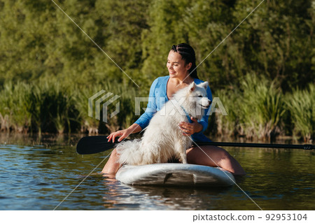 Happy Young Woman on the Lake at Early Morning Sitting on the Sup Board with Her Dog Snow-White Japanese Spitz and Enjoying Sunrise Happy Young Woman on the Lake at Early Morning Sitting on the Sup Board with Her Dog Snow-White Japanese Spitz and Enjoying Sunrise 92953104