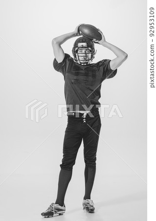 Portrait of young man, american football player posing in helmet and uniform with ball. Black and white photography Portrait of young man, american football player posing in helmet and uniform with ball. Black and white photography 92953109