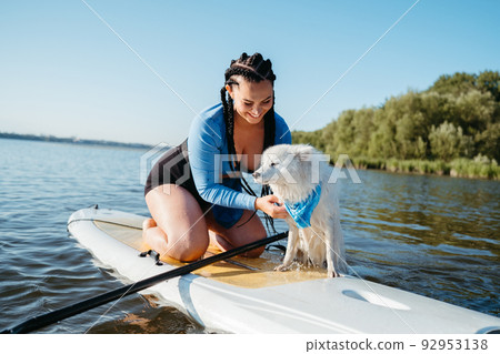 Cheerful Woman with Locs Helping Her Dog Japanese Spitz Get Out of the Water, While Sitting on the Sup Board on the Lake 92953138