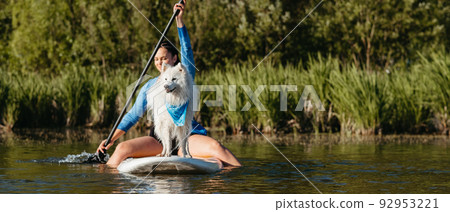 Young Woman Paddleboarding with Her Pet on the Lake, Snow-White Dog Breed Japanese Spitz Standing on the Sup Board 92953221