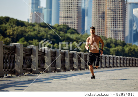Urban sports. Sporty man shirtless jogging for fitness in the city park on a beautiful summer day 92954015