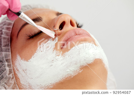 Satisfied young Caucasian beautiful woman uses services of professional beautician in beauty salon or spa. Unrecognizable beautician applied white mask to half of client's face with brush. Top view. 92954087