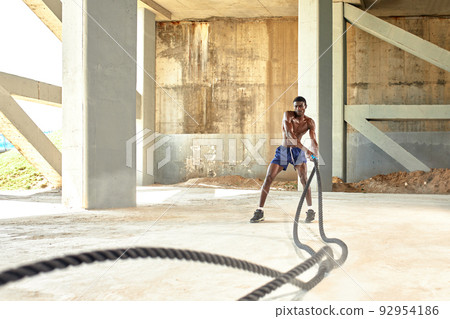 Rope workout. Sport man doing battle ropes exercise outdoor. Black male athlete exercising, doing functional fitness training with heavy rope 92954186