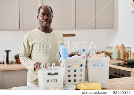 Portrait of African mature man looking at camera while separating plastic, paper and glass in different containers in kitchen 92955575