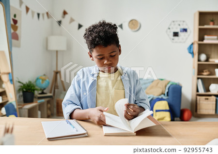 African little boy turning the pages of his book at the table while doing his homework at home 92955743
