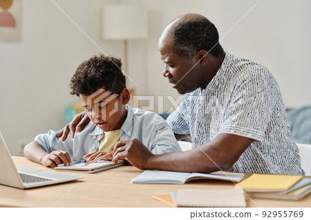 African teacher explaining new material to schoolboy pointing at notebook while they sitting at desk in the room African teacher explaining new material to schoolboy pointing at notebook while they sitting at desk in the room 92955769