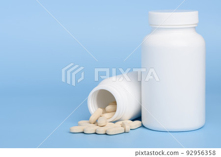Two white medicine bottles, one closed and second with pills spilled on blue background  92956358