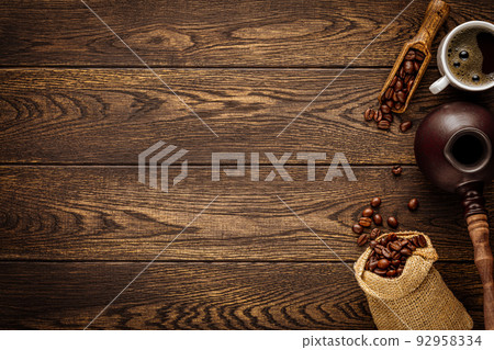 Natural turkish coffee. Freshly brewed aromatic drink in cup, roasted beans, vintage cezve pot and wooden spoon, burlap sack bag over rustic wood background. Copped image with copy space Natural turkish coffee. Freshly brewed aromatic drink in cup, roasted beans, vintage cezve pot and wooden spoon, burlap sack bag over rustic wood background. Copped image with copy space 92958334
