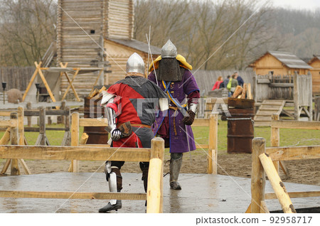 Men reenactors in metal armor of an Old Rus knights reconstructing sword fight, wooden fortress on a background 92958717