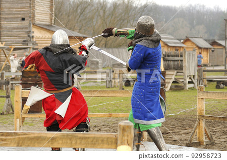 Men reenactors in metal armor of an Old Rus knights reconstructing sword fight, wooden fortress on a background Men reenactors in metal armor of an Old Rus knights reconstructing sword fight, wooden fortress on a background 92958723