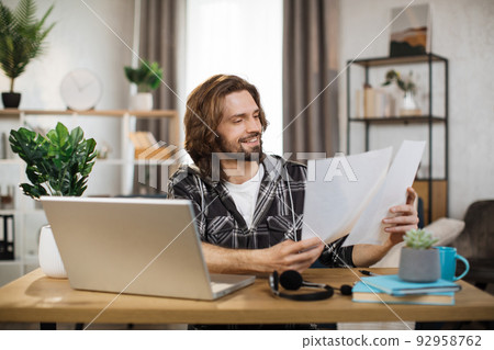 Likable young caucasian man sitting at desk with laptop and holding papers. 92958762