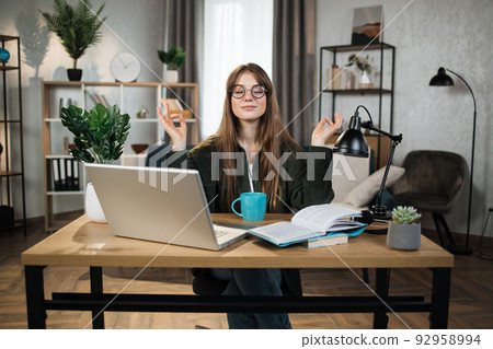 Woman freelancer in eyeglasses sitting at table with closed eyes and relieving stress by meditation Woman freelancer in eyeglasses sitting at table with closed eyes and relieving stress by meditation 92958994