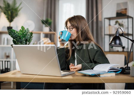 Attractive woman using modern laptop while sitting at desk and drinking hot coffee. Attractive woman using modern laptop while sitting at desk and drinking hot coffee. 92959060