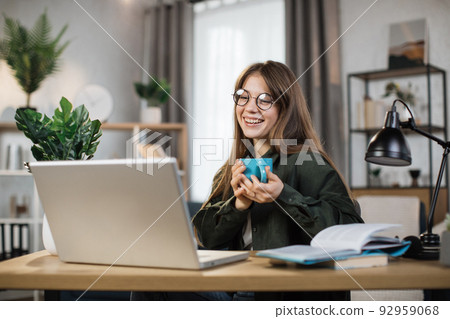 Excited young woman holding cup with hot drink and laptop for video conversation Excited young woman holding cup with hot drink and laptop for video conversation 92959068