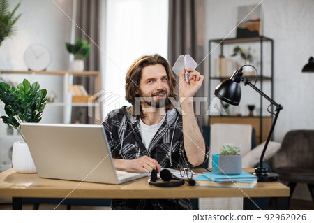 Caucasian man in casual outfit sitting at desk with modern laptop and playing with paper plane. 92962026