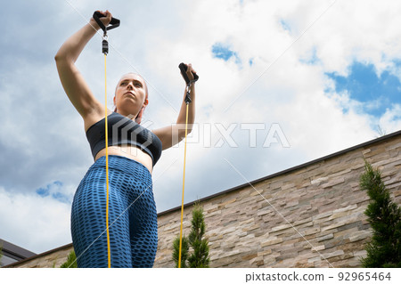 Attractive young fitness girl practising stretching exercise with rubber bands outdoors in the garden of her house. health and wellness concept. Attractive young fitness girl practising stretching exercise with rubber bands outdoors in the garden of her house. health and wellness concept. 92965464