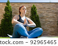 young woman with red hair, practising yoga in the garden in the open air, relaxation exercises, doing the lotus posture. concept of health and well-being. 92965465