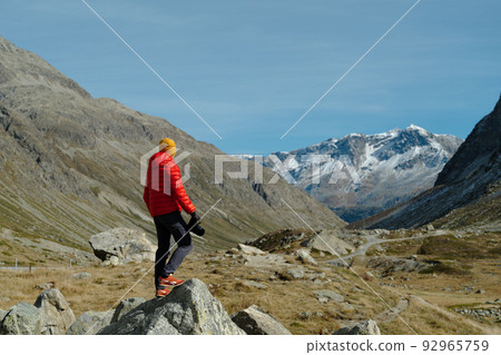Back view of a tourist man in red jacket and camera standing with a view on Julier Pass, Switzerland Back view of a tourist man in red jacket and camera standing with a view on Julier Pass, Switzerland 92965759