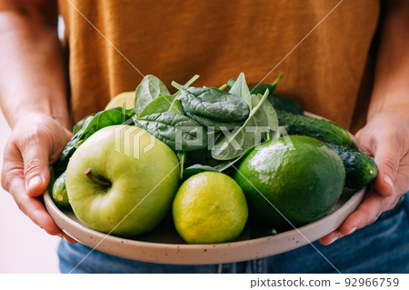 A woman holds in her hands fresh green vegetables and fruits in a large plate, close-up soft focus 92966759