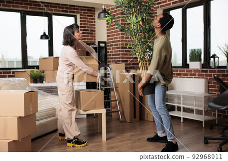 Diverse couple buying first home and moving in together, unpacking plant and furniture to decorate new household. Enjoying property relocation with decor to start new lifestyle beginnings. 92968911