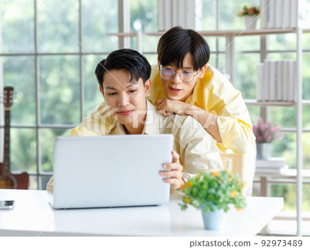 Two Asian young happy pride teenager male gay men lover couple partner sitting smiling hugging cuddling together in front laptop notebook computer browsing surfing using credit card shopping online 92973489