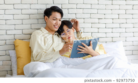 Two Asian young happy romantic teenager male gay men lover couple partner laying down under blanket on bed in bedroom leaning at brick wall background cuddling hugging together while reading novel 92973750