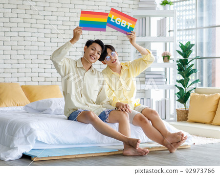 Portrait shot Two Asian young happy cheerful teenager male gay men lover couple partner sitting holding hands together on bed in bedroom showing rainbow LGBTQ card board celebrating June pride month Portrait shot Two Asian young happy cheerful teenager male gay men lover couple partner sitting holding hands together on bed in bedroom showing rainbow LGBTQ card board celebrating June pride month 92973766