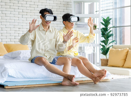 Two Asian young happy playful teenager male gay men lover couple partner sit on bed in modern bedroom wearing virtual reality VR goggles headset having fun playing online 3D simulator game together Two Asian young happy playful teenager male gay men lover couple partner sit on bed in modern bedroom wearing virtual reality VR goggles headset having fun playing online 3D simulator game together 92973785