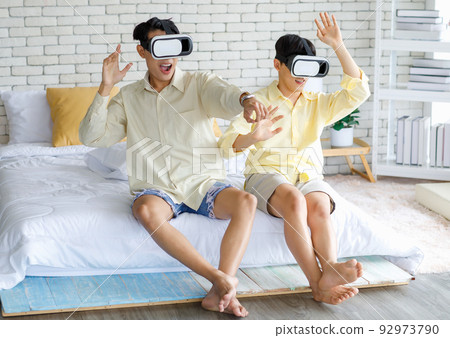 Two Asian young happy playful teenager male gay men lover couple partner sit on bed in modern bedroom wearing virtual reality VR goggles headset having fun playing online 3D simulator game together 92973790